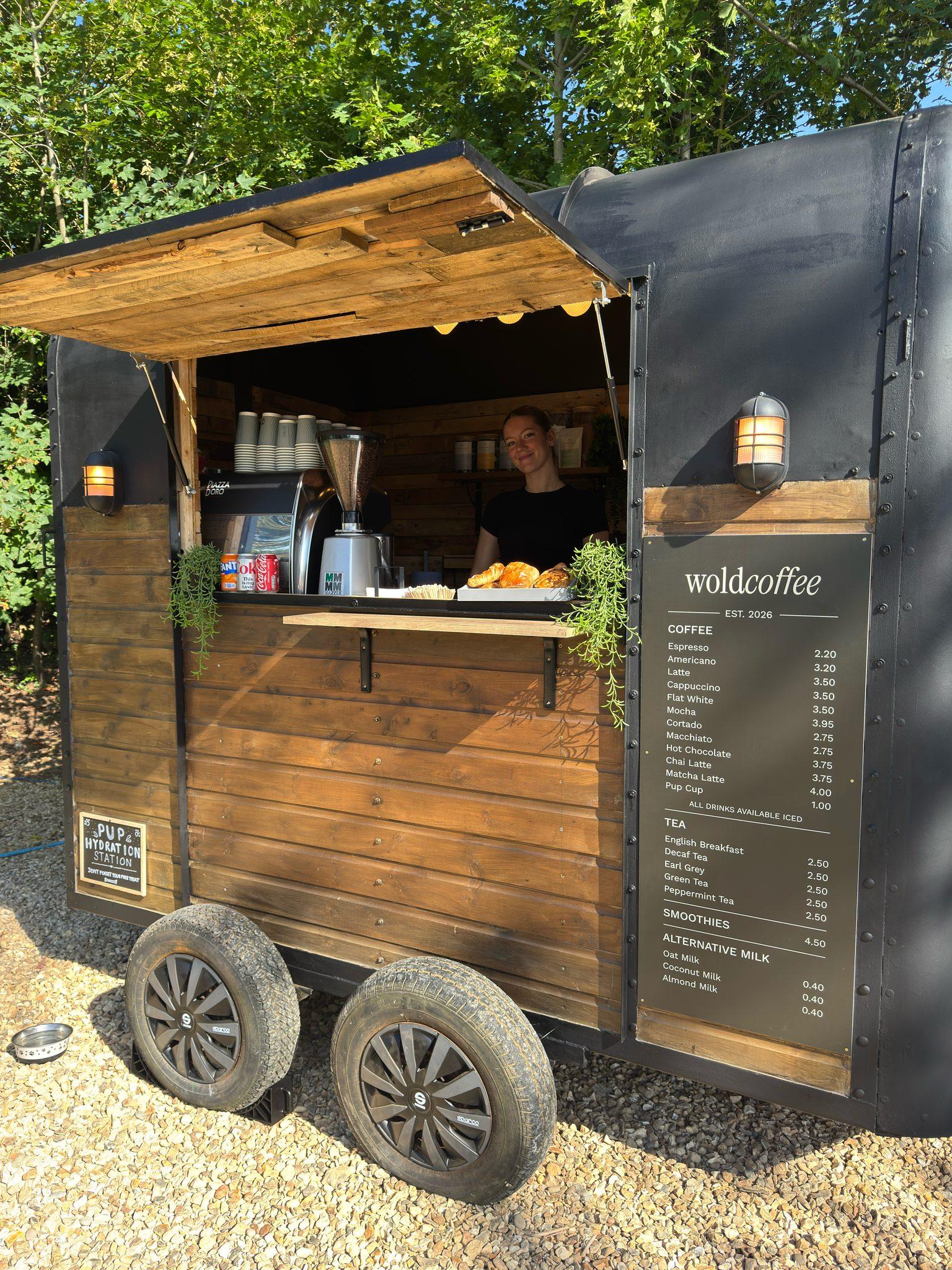 The WoldCoffee weekday trailer, a converted horsebox clad in wood under a black roof, its hatch propped open on a gravel pitch with trees behind. A barista stands at the counter beside a tray of pastries, and the menu board is fixed to the side panel.